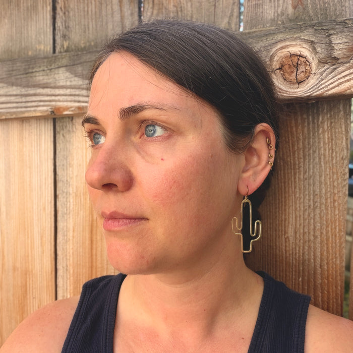 A woman with dark hair pulled back, wearing a black tank top and a gold cactus-shaped hoop earring, photographed outdoors against a rustic wooden fence.
